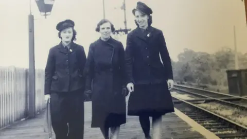 Three women stand on a train platform. The photo is in black and white. To the right of the photograph is the trail track, to the left is an old fashioned street lamp. Two of the women are wearing their railway issue hats. All are in dark, 1940s-style clothing