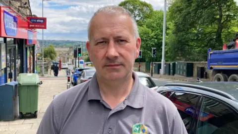 A man in a grey polo top standing on a pavement with a road behind him.