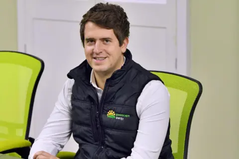 Josh Young Josh Young sits at a table and smiles at the camera. He has short dark hair and wears a white shirt with a black gilet jacket over the top.