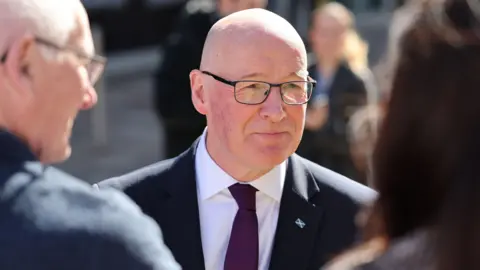 PA Media John Swinney, who is bald with glasses, wearing a business suit and a purple tie. He is facing two people, out of focus in the foreground 