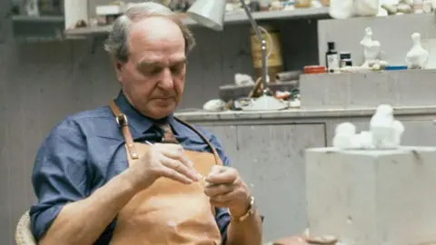 Getty Images The artist Henry Moore in his studio, he is wearing a leather apron with a blue shirt underneath and has grey hair swept over his head. There are various artists' materials in the background.