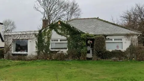 Google A white-rendered bungalow, with a grey tiled roof and ivy growing around one window and the door, sits on a plot of green grass