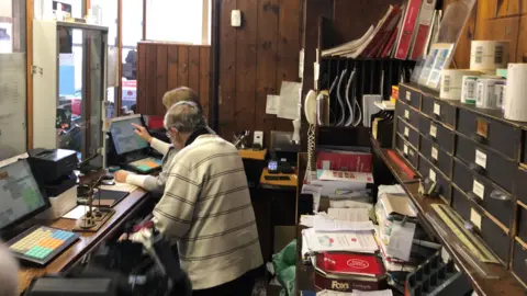 A woman touching a computer screen behind a counter with a man standing behind her. It is a busy scene with bundles of papers, sorting boxes, a wooden counter and computers. 