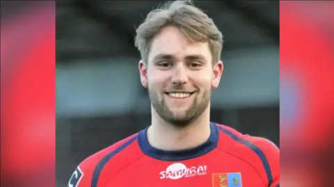 Jon Devereux is seen from his shoulders up wearing a red rugby uniform. He is smiling.