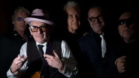 Ray Di Pietro Elvis Costello and The Imposters with Charlie Sexton posing for a publicity image.