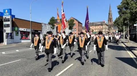 Members of the Orange Order marching. They are each dressed in grey trousers, black coats, white gloves and orange sashes. The have black top hats on. The sky is cloudless and blue.
