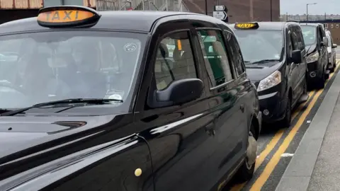 BBC A row of black taxis with their lights on, parked on a double yellow line on the side of the road.