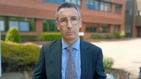 Muir is wearing a navy suit with a blue shirt and a blue tie that's patterned with a gold design. He wears glasses. A building is behind him.