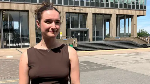 Sophie Watson wearing a sleeveless top and jeans stands on a paved area outside a modern office building with large glass windows and a concrete façade, with steps and the building entrance visible in the background under bright sunlight.