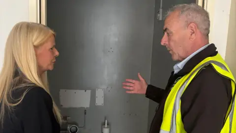 South Ribble Borough Council handout Councillor Clare Hunter with long blond hair wearing a black blazer talks to a builder with grey hair and wearing a hi-vis vest over a black long-sleeved top. They are standing in front of a bathroom being refurbished. The image shows them from the side.