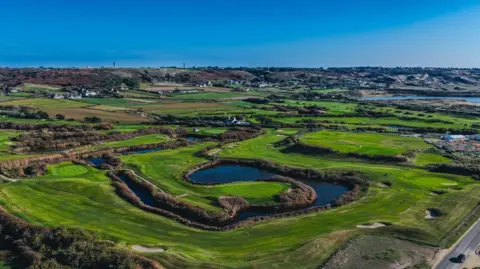 Craig Robinson/ Savills A green golf course is besides the sea and has a large pool of water in the centre.