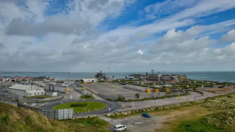 Getty Images A view of Rosslare port in Co Wexford. In view there is a roundabout with green grass in the middle. There is also a car park and general buildings. In the distance the sea is visible. To the front of the image there is a rough area of grass and dry ground.  