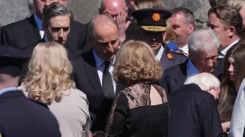PA Media A crowd of people in dark suits, a bald man speaks to a woman with blonde, curly hair, her back is facing the camera
