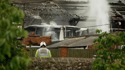A firefighter in a high-visibility jacket and helmet stands behind a low stone wall, facing a heavily damaged building where water sprays over a charred roof and rising steam. The structure has exposed beams, broken roofing, and red brick walls, with fencing and greenery partially framing the foreground.
