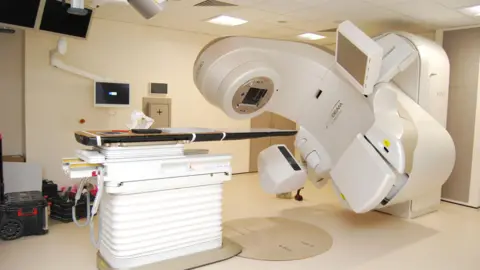 A view of a linear accelerator, a large white robotic, C-shaped machine that sits in a specialized, shielded treatment room. It consists of a tall, stationary stand (or "stand") and a rotating, horizontal arm called a gantry, which houses the radiation beam source