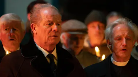 BBC/World Productions Peter Mullan in a suit standing next to a vicar with people holding candles