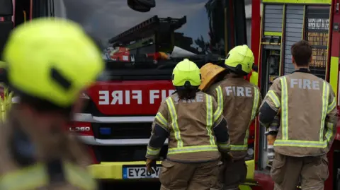 Three firefighters walk towards a fire engine with another crew member blurred in the foreground. They wear brown uniforms with fluorescent strips, with 'fire' printed on the uniforms in big white letters.