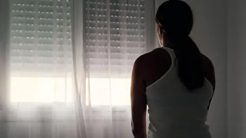 Getty Images A stock image of a woman sitting on a bed staring out of a window covered by a net curtain.