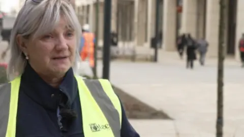 A woman with shoulder-length grey hair. She is wearing a black top with a yellow hi-vis jacket on top.