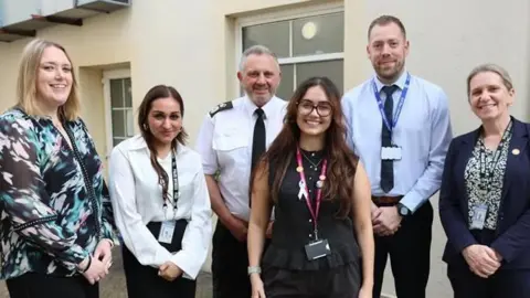 Guernsey Police The image shows seven people standing together outdoors, arranged in a loose group in front of a light‑colored building. They are all dressed in professional or business attire, and each person is wearing an ID badge on a lanyard.