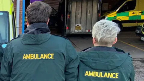 A man and woman have their backs to the camera and are facing an ambulance station. The man has short black hair and the woman has short bleached blond hair. They are both wearing green hooded coats with the word ambulance written in yellow across their back