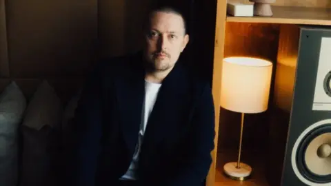 Hallwood Media A man is sitting on a dark coloured sofa that has grey pillows on it. He has white skin and is wearing a white t-shirt and navy blazer. To the right of the image there is a yellow lamp and large black boom speaker. 