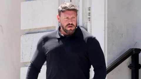Karl coming out of court. He has short brown hair and a groomed moustache and beard. He has a black top on and grey jeans. he holds his phone in his left hand.