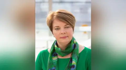 Suffolk County Council This is a professional headshot of Nicola Beach, She is wearing a peacock green jumper with a matching coloured scarf with peacock feathers on it. She is smiling at the camera 