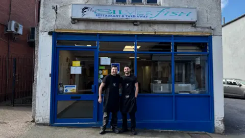 Two men with dark hair in dark aprons, dark tshirts and dark trousers outside the front of a shop with blue doors and window frames and a sign above the door which reads Silverfish