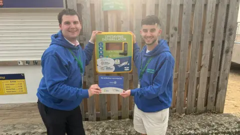 Donate For Defib Two men in blue hoodies standing next to a defib.