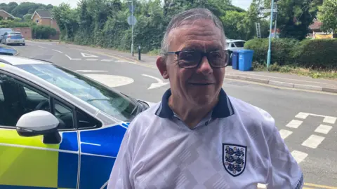 An older man with grey hair wears an England football shirt and glasses. He is standing on a pavement in front of a mini roundabout and a police car is parked behind him.