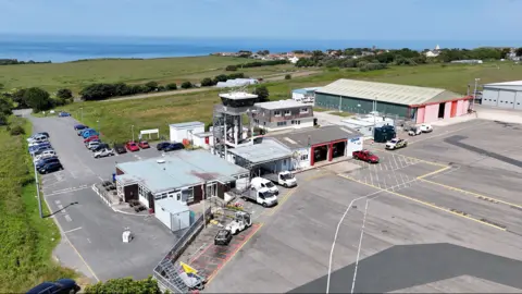 A drone image taken from above of Alderney Airport. It is a large building with a number of vehicles parked next to the runway.