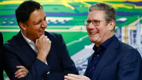 Getty Images Anas Sarwar and Keir Starmer stand side-by-side, left and right of shot. The backdrop appears to be a very colourful technical graphic. Both men are in blue suits, Sarwar has a white shirt, Starmer has a dark blue one. They are not wearing ties and are laughing together.