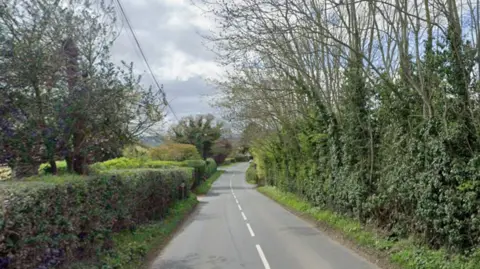 Google The image shows a long two-lane country road with trees on either side.