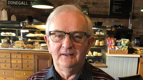 Life After Bobby Bradley in a cafe, he has short white hair and is wearing black framed glasses. He has a striped maroon jumper on with a blue and white pinstriped shirt. 