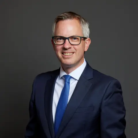 UK Parliament A studio portrait of a man wearing a dark blue suit, white shirt, blue tie, and glasses, standing against a dark gradient background.