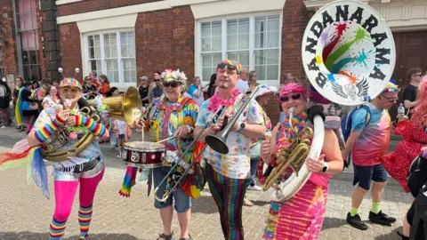 Four people in as much colourful clothing as they can get on, stand with brass instruments. Bandanarama is written in the tuba.