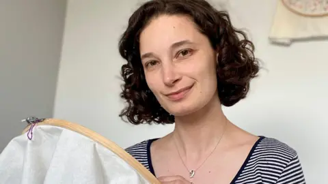 Sophie O'Neill A woman with a short bob and a stripy black and white t-shirt smiling at the camera while holding her embroidery hoop.