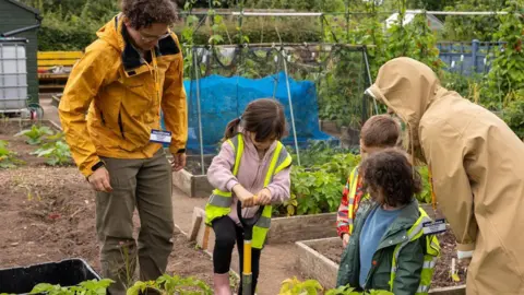 Earlsdon Primary School Three children at the plot. All are wearing hi-visibility green jackets and a girl is using a spade. A man and a woman are supervising and plants can be seen growing. 