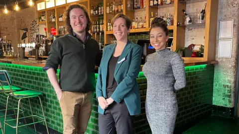 A man and two women stand in front of a bar. The man on the left has chin-length brown curly hair and is wearing a grey jumper and brown trousers. The woman in the centre has short brown hair and is wearing a dark green suit jacket. The woman on the right has long brown hair tied back and is wearing a grey dress. The bar has green tiles and stools.