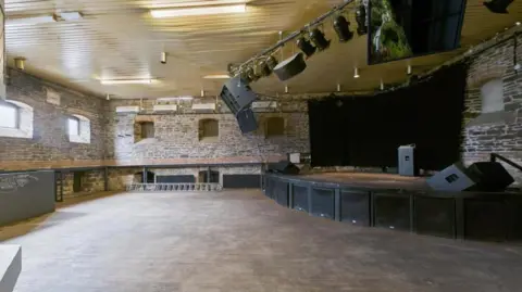 Hollis and Morgan An empty music venue. There is a small stage to the right, with a black curtain covering a stone wall and a lighting rig above it. There is a wooden dancefloor filling the rest of the room.