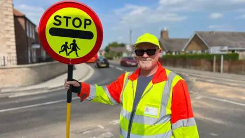 Neil Cotton dressed in his school crossing patrol hi-vis clothing. He is standing in a road holding up his stop sign. 