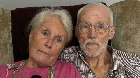 A woman and a man are seated side by side on a cream-coloured chair, which has a blue covering draped over it. The woman is wearing a pink top and an apron, has long grey hair, and is staring directly at the camera. The man is dressed in a red and blue checked shirt, unbuttoned at the top, and has short grey hair, a grey beard and glasses. Both individuals appear upset.