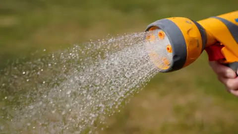 A hosepipe being used in a garden. Water is sprinkling from the yellow and grey hose attachment with green grass in the background.