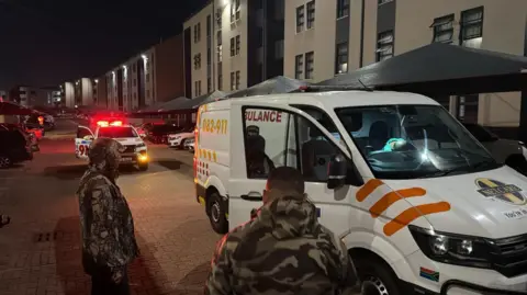 South Africa Police Service Men in camourflage jackets stand by an ambulance in a residential area.