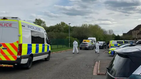 Several police vehicles in a road with two forensic officers, dressed in white, walking away. There are other cars parked to the right and houses to the right. A park area is to the left with grass and lots of trees. A lamp post is to the right. 