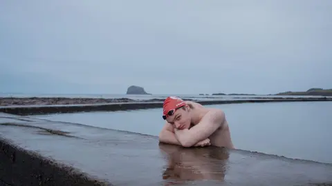 Jennifer Charlton Male swimmer with red swim cap leans on concrete at side of outdoor pool, looking at camera, against grey background.