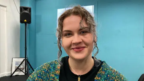 A young woman with curly hair, wearing a black T-shirt and a green, yellow and blue cardigan.