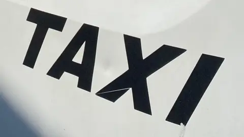 BBC Black writing which reads TAXI written on a white metallic side of a private hire taxi