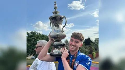 A man with brown hair swept over his forehead smiling and holding up a large silver trophy. He is wearing a blue Scunthorpe United top.